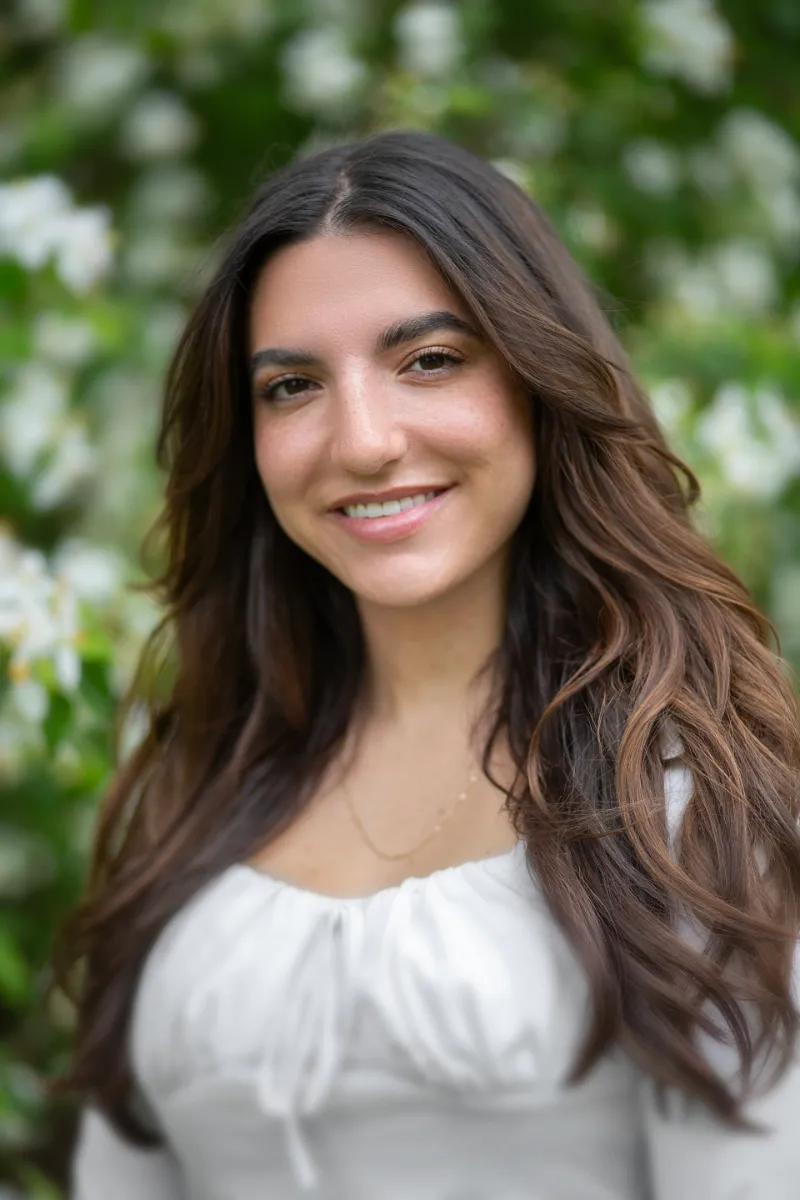 Outdoor headshot of a young woman with wavy brown hair smiling among white blossoms