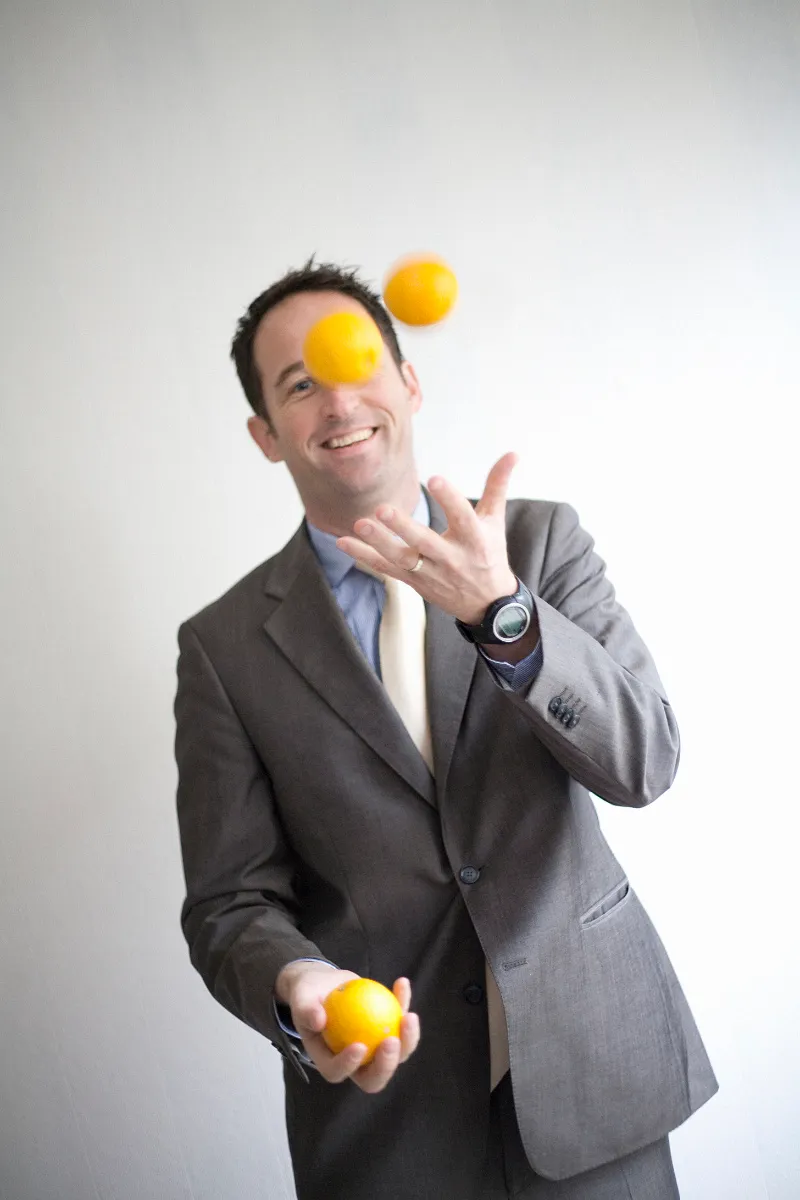 Playful business portrait of a man in a grey suit juggling oranges with a bright smile