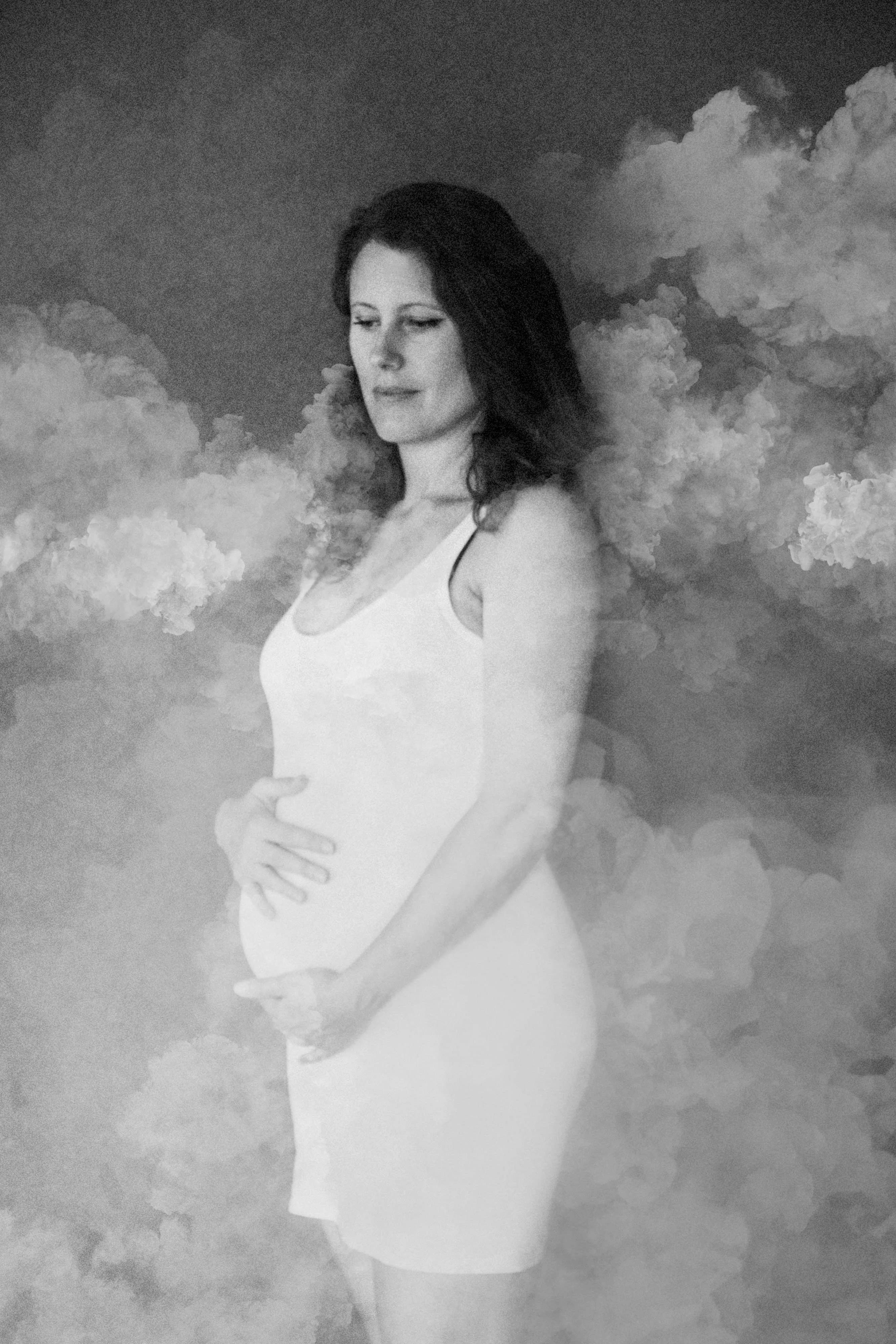Black and white maternity portrait of a pregnant woman cradling her belly among soft clouds