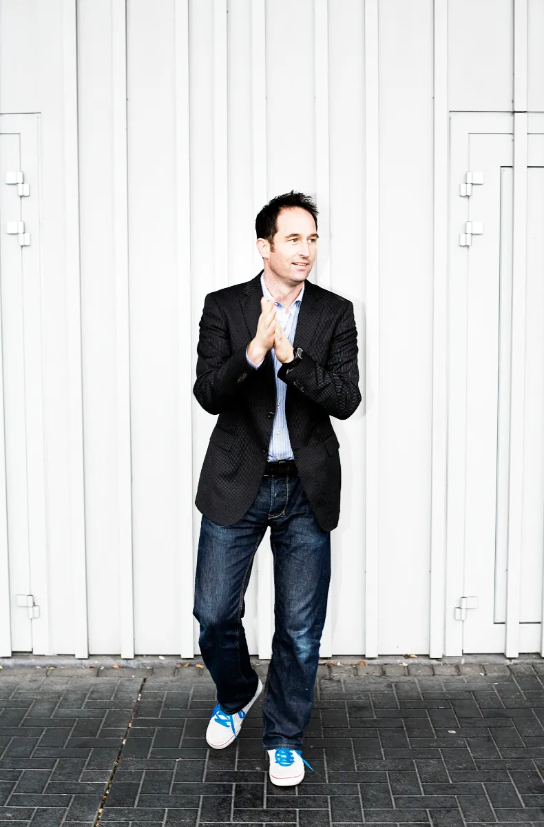 Casual business portrait of a man in a dark blazer and jeans with hands clasped against a white wall
