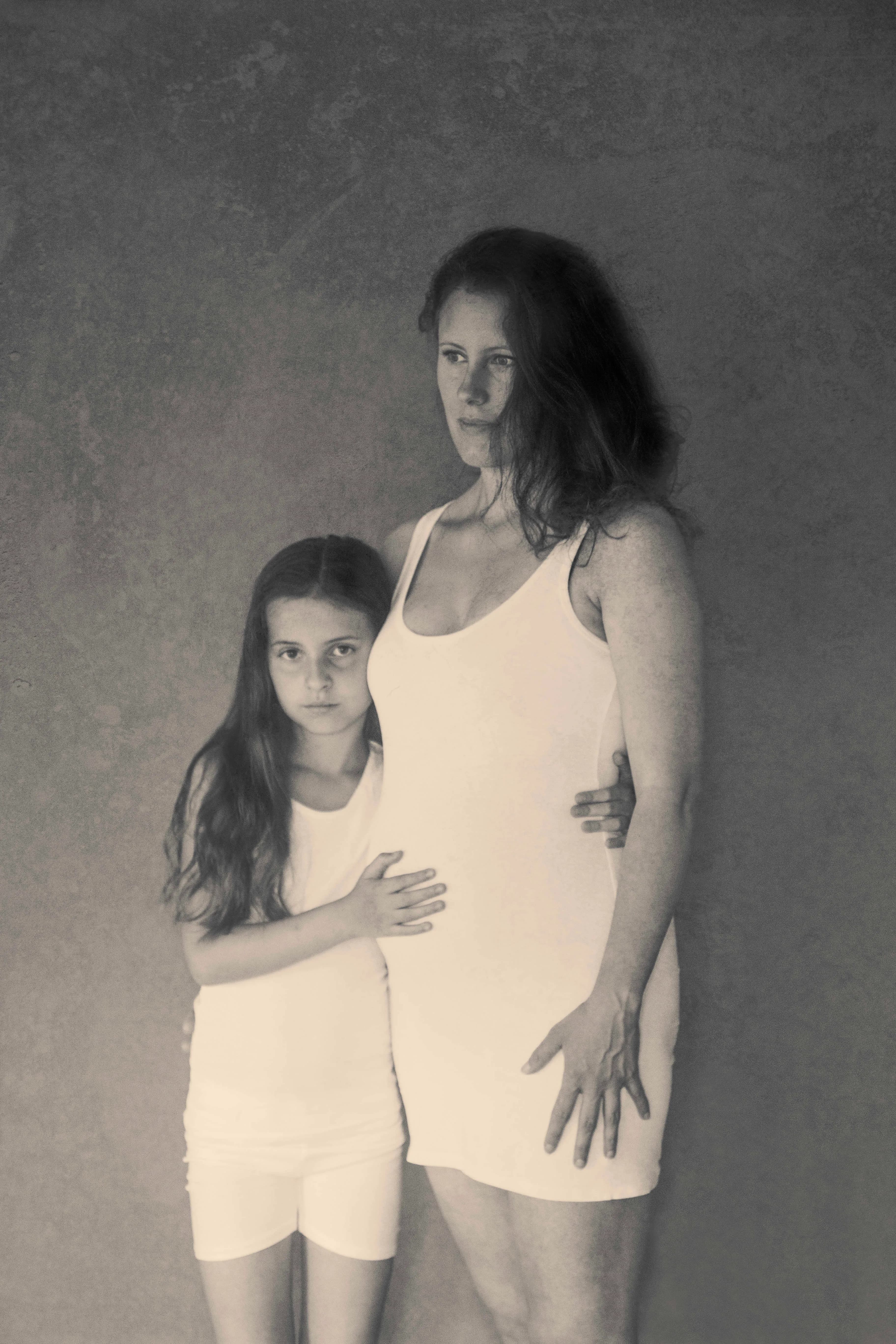 Soft toned maternity portrait of a pregnant mother with daughter holding her belly in studio